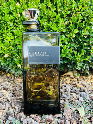 一瓶 Corzo 龍舌蘭酒放在木屑上，後面有綠色灌木叢。