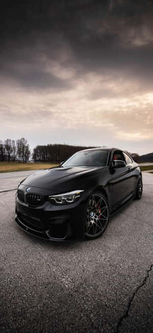 黑色BMW M2雙門跑車停在裂開的瀝青路上，背景是暴風雨黃昏的天空。 — iPhone 17e 壁紙