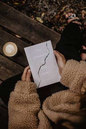 一個人在秋天戶外喝咖啡時閱讀名為《The Strength in Our Scars》的書。