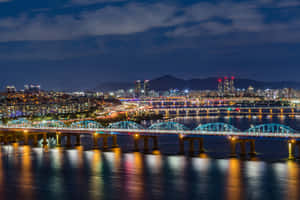 燈火通明的橋樑和城市燈光在水面反射，背景有山脈的夜景。 — MacBook 壁紙