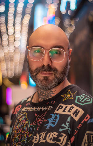 一個光頭、戴眼鏡、留鬍鬚的男人，穿著圖案T恤，身處霓虹燈照亮的夜晚城市。