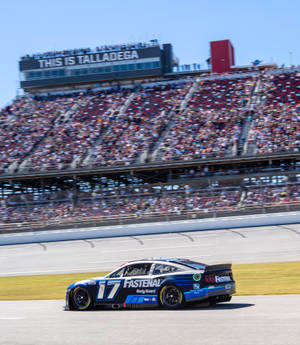 藍黑相間的NASCAR賽車在賽道上高速行駛，背景是歡呼的人群。
