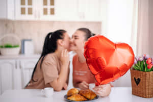 兩位女士在廚房裡接吻，旁邊有紅色心形氣球和牛角包。
