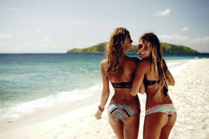 兩位穿著彩色比堅尼的女性在陽光普照的海灘上擁抱，旁邊是碧綠的海水。 — 海灘 壁紙