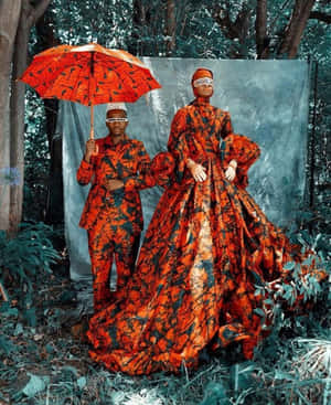 兩個穿著鮮豔橙色圖案服裝的人在森林中撐著雨傘擺姿勢。 — 黑色 壁紙