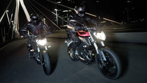 兩名車手駕駛跑車在夜間燈火通明的橋上飛馳。 — 機車 壁紙