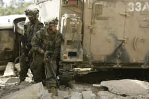 兩名身穿迷彩裝備的武裝士兵站在一輛軍用卡車旁，身處瓦礫遍地的區域。 — MacBook Air 15 壁紙