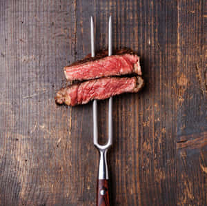 兩塊 medium rare 牛扒片喺金屬叉上，襯住深色木質背景。 — 1:1 壁紙