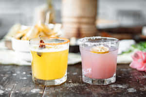 木桌上兩杯色彩繽紛的雞尾酒，配以小吃和花朵裝飾。 — MacBook Pro 14 壁紙