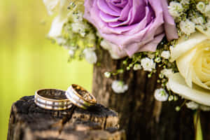兩枚刻有圖案的結婚戒指放在樹樁上，伴隨著紫色和白色的玫瑰。 — MacBook Pro 壁紙
