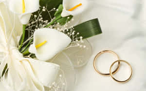 兩枚金色結婚戒指旁邊有一束附有絲帶的白色馬蹄蓮。 — 1280x800 壁紙