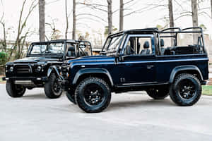 兩輛黑色 Land Rover Defender 停泊在戶外樹木附近。 — Surface Pro 壁紙
