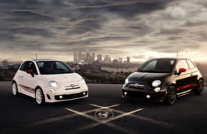 日落時分，兩輛Fiat 500汽車停在瀝青路上，背景是城市天際線。 — Surface Pro 壁紙