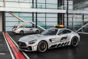 兩輛銀色 Mercedes-AMG F1 安全車，閃著燈，停在一棟現代玻璃建築前。 — Surface Pro 壁紙