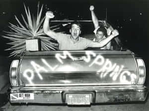 夜晚，兩名男子在尾門上噴有「PALM SPRINGS」的皮卡車中慶祝。 — 70年代gif 壁紙
