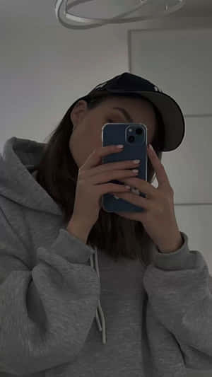 穿著灰色連帽衫和帽子的人在室內用藍色手機拍鏡子自拍。 — 美學 iPhone 壁紙