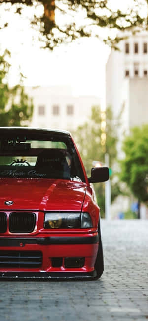 紅色BMW E30停在鵝卵石街道上，樹木和建築物柔和對焦。 — Pixel 8a 壁紙