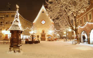 夜晚白雪覆蓋的小鎮廣場，建築物燈火通明，節日燈光閃爍。
