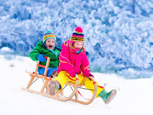 兩個孩子穿著冬季裝備，在雪坡上滑雪橇時大笑。 — 雪 壁紙