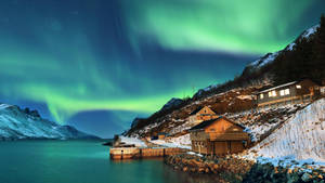 北極光在積雪的山脈和寧靜湖邊的小屋上空發出綠光。