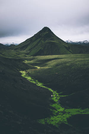 一條綠色河流流經深色火山山谷，上方是多雲的山峰。