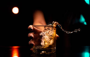 手將琥珀色飲料連冰塊倒入玻璃杯，在彩色燈光下