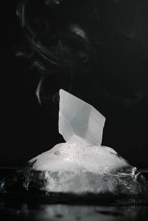 一顆從融冰中升起的晶體，煙霧在其上方旋轉，背景為深色表面。