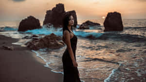 身穿黑色連衣裙的女性在日落時站在海灘上，有海浪和岩石。
