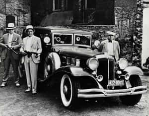 三名身穿西裝和帽子的男子站在一輛1930年代復古汽車旁，背景是磚砌建築。