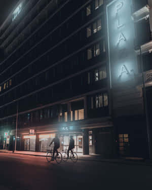 兩名騎單車的人在夜間沿著城市街道騎行，經過一座有垂直招牌的明亮建築物。