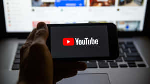 手拿著帶有YouTube標誌的手機，前面是筆記本電腦 — YouTube 壁紙
