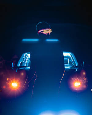戴著發光眼鏡的人站在一輛車頭燈明亮的汽車前，夜晚。 — 霓虹 iPhone 壁紙