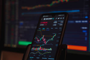 智能手機顯示加密貨幣交易圖表，背景有電腦顯示器