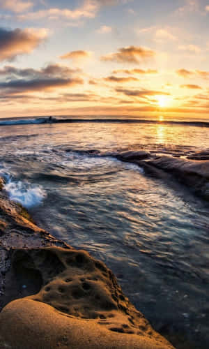 海上夕陽，海浪拍打岩石，遠處有衝浪者。