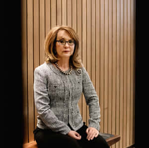 一位戴眼鏡、身穿質感外套的女士坐在室內木長椅上。 — 政治 壁紙