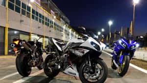 三輛Yamaha跑車在夜間停泊在賽車設施外，在明亮燈光下。 — iMac 壁紙