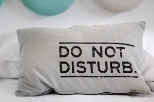 灰色枕頭，上面有「DO NOT DISTURB」字樣，放在一張鋪有白色床單的床上，背景有青綠色氣球。