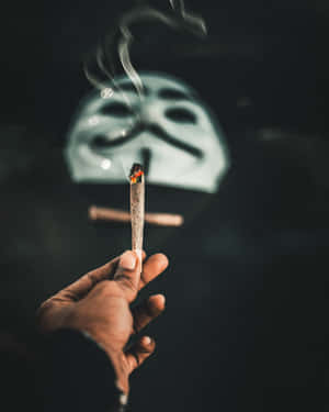 手拿著點燃的大麻煙，煙霧升起，背景是模糊的蓋伊·福克斯面具。 — 食草 壁紙