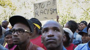 抗議者在戶外聚集，在樹下舉著「STOP RACISM」標語牌。