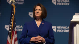 身穿藍色西裝的女性在政治活動中，背景有美國國旗和Nevada Victory標誌。 — 政治 壁紙