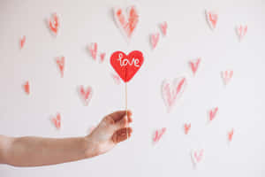 手拿著一個紅色心形，上面寫著 love，附在棍子上，對著一面裝飾著心形的牆壁。