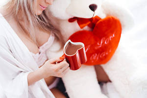 身穿白色浴袍的女性，手持紅色心形杯，旁邊有泰迪熊和「I Love You」枕頭。