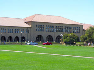 一座大型大學建築，帶有拱形窗戶和赤陶屋頂，位於綠色草坪後面，草坪上停有汽車和行人。 — 建築 壁紙