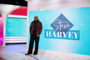Steve Harvey 在工作室微笑，身後藍色屏幕上顯示他的名字。