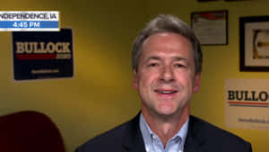 穿西裝的男子在競選辦公室微笑，旁邊有Bullock 2020標誌。 — 政治 壁紙