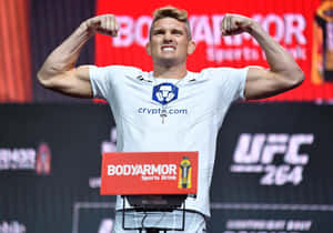UFC 拳手在稱重活動中彎曲二頭肌，身穿帶有加密貨幣標誌的白襯衫。