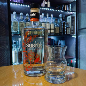一個Suerte龍舌蘭酒樽同空杯喺吧枱上，後面有發光嘅架。