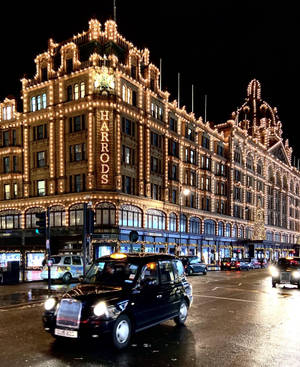 黑色出租車在濕滑街道上駛過夜晚燈火通明的Harrods百貨公司。 — Galaxy Z Fold 5 壁紙