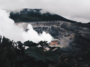 火山口噴出蒸氣，前景有藍綠色水和茂盛的綠色植物。