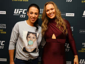 兩位女性在UFC活動中，一位穿著休閒T恤，另一位穿著優雅連衣裙，兩人都微笑。